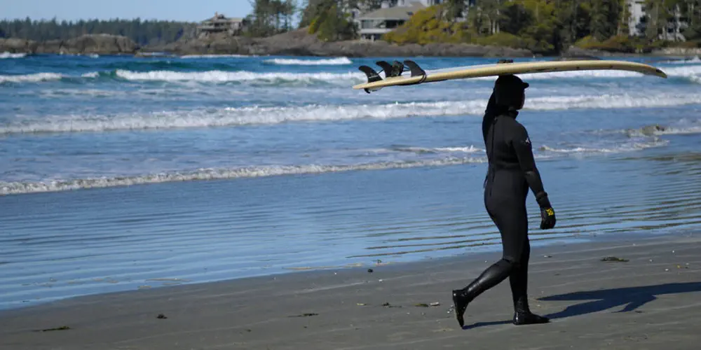 A female surfer in a wet suit walks on the shore carrying a surfboard over her head.