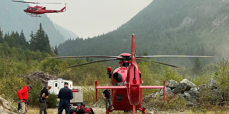 One red helicopter sits on the ground while people get ready to fly it. Another red helicopter has just taken off.