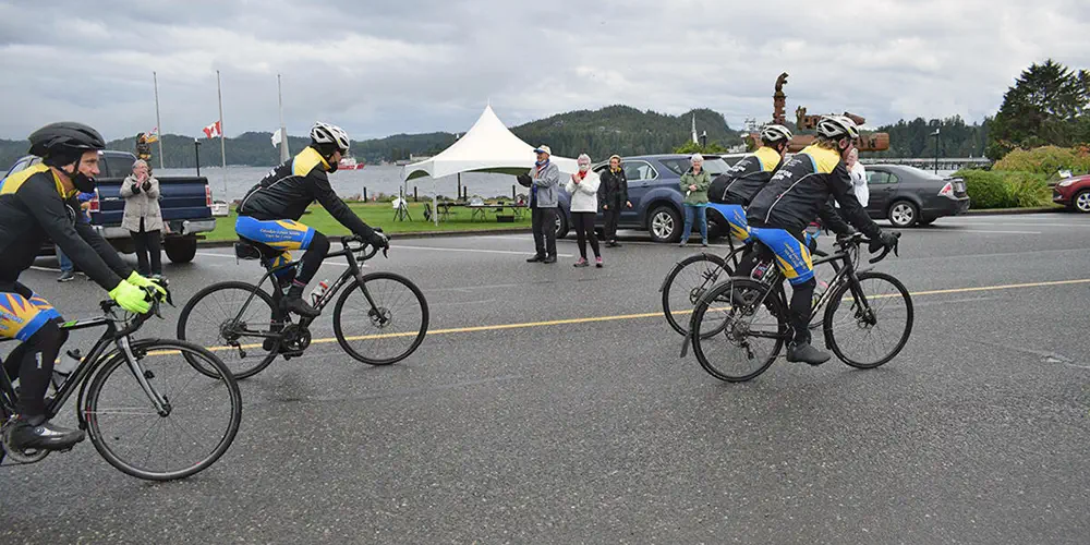 Cyclists ride off while people wave on a cloudy day.