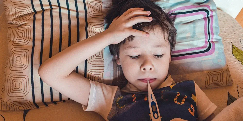 A little kid lays on a pillow with his eyes closed. He has his hand on his forehead and a thermometer in his mouth.