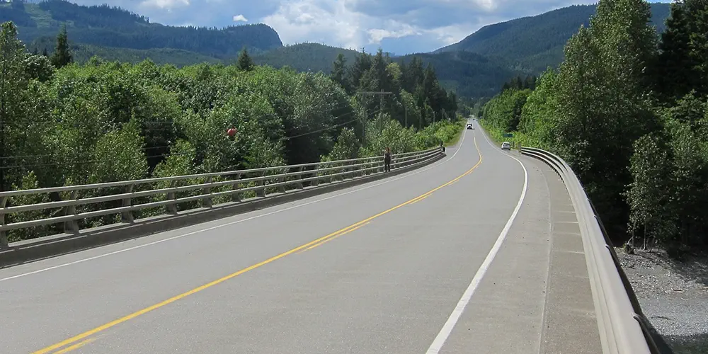 A picture of Highway 19 going off into the distance on a sunny day. One person is driving, another is walking on the sidewalk.