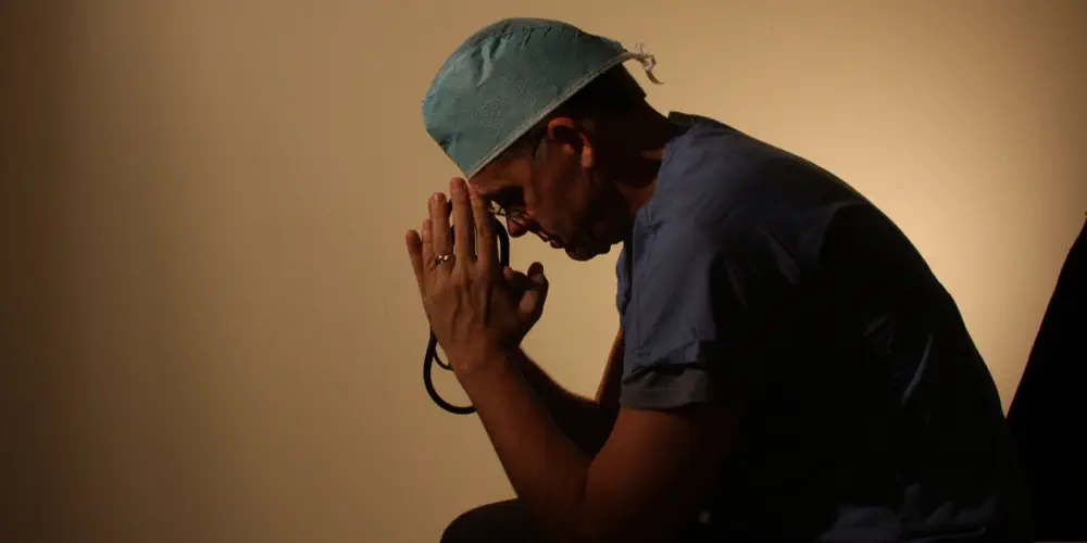 A doctor sits in a dark room with his head in his hands.