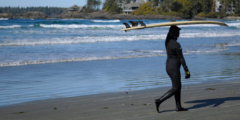 A female surfer in a wet suit walks on the shore carrying a surfboard over her head.