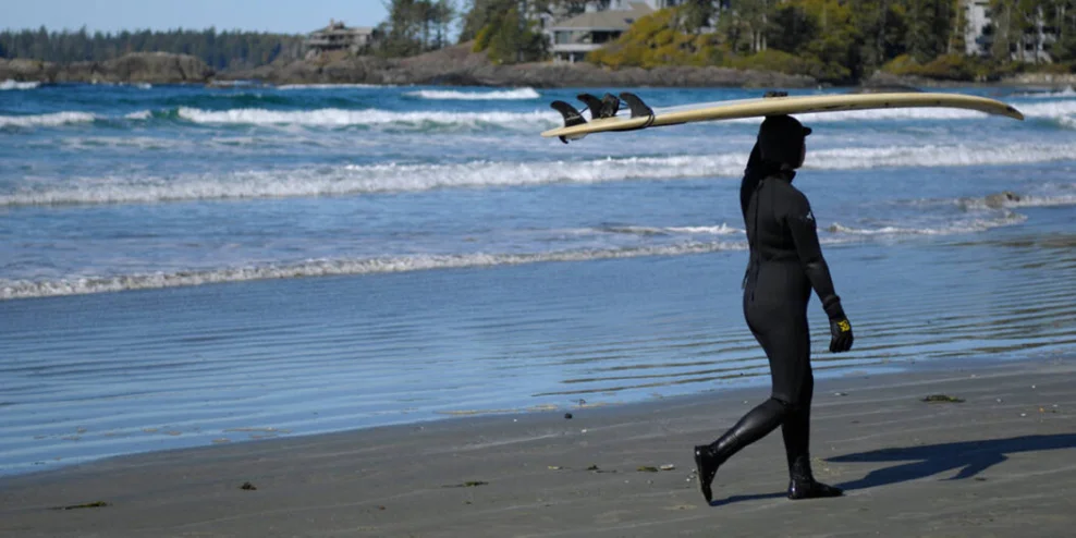 A female surfer in a wet suit walks on the shore carrying a surfboard over her head.