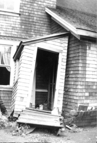 A black and white closeup of the side of a house where the entryway has come away from the building and is leaning.