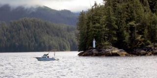 A small boat sits in the Muchalat Inlet in front a rocky shore and tall trees. It is cloudy.