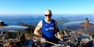 Jesse Barnes plays his drums with a sea and mountain vista in the background. It's sunny and he is wearing sunglasses.