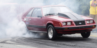 An old red sports car takes off from the starting line and leaves smoke behind the tires.