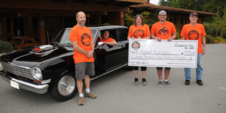 A group of smiling people stands in front of a shiny black car while holding up a huge novelty cheque.