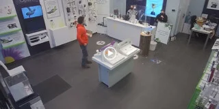 A man in a red hoodie stands in the middle of the cell phone store with a carton of eggs. Two employees sit behind the desk looking bewildered.
