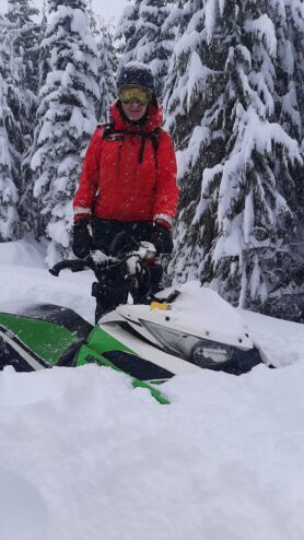 A member of the Comox Valley SAR stands with a snowmobile. It is nearly buried in snow.