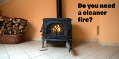 A wood stove sits against a peach coloured wall. There's a neat pile of firewood beside it.