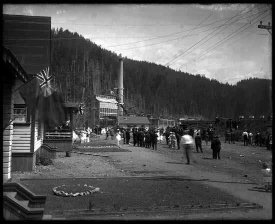 A black-and-white photo of people gathering in Port Alice in the early 1900s. The smoke stack from the old mill is in the background.