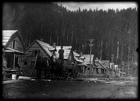 A black-and-white photo of a man with a horse and buggy in front of houses being built. Tall trees stand in the background.