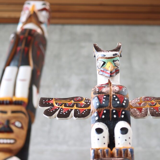 A close-up of the top part of Judith Sayers' new talking stick in front of a much larger totem pole.