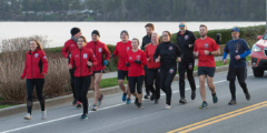 A group of runners in red shirts and black bottoms run along a road next to a lake. The weather is not very good.