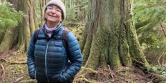 A woman in a blue puffy jacket and knit toque smiles in front of a huge mossy tree.