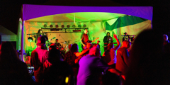 A band plays on an outdoor stage at night with colourful lights on the stage. The lead singer looks very stoked to be there.