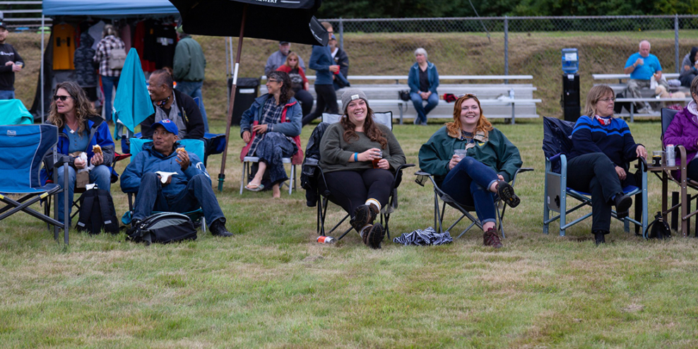 A group of people sit on lawn chairs on the grass at an outdoor field. They look comfortable and happy and are drinking what look to be beers.