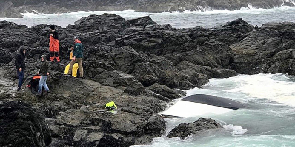 Three men and one of their rescuers stand on a rocky outcropping in stormy seas. Their boat is upside down in the water.