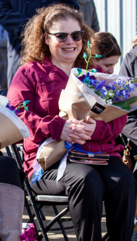 Melissa Fletcher sits in a chair at an outdoor ceremony. She's carrying a bouquet of purple flowers that match her pink coat. Her smile is huge.