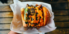 A closeup of a hand holding a loaded burger in paper packaging. The toppings are outrageous.