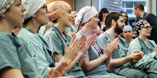 A room full of medical school students claps for a colleague on a stage that sits out of frame.