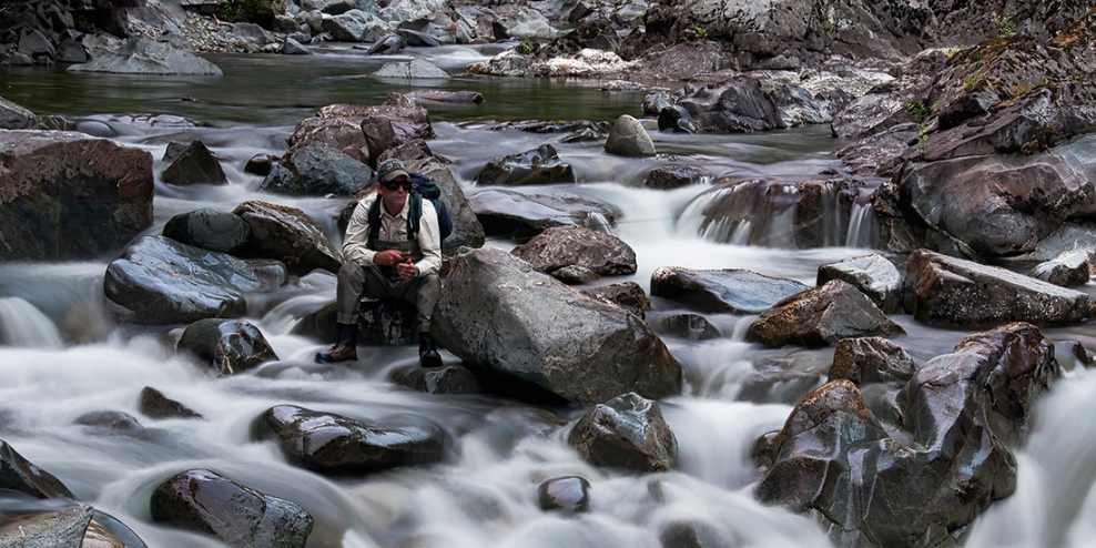 A fisher sits on a rock in a river and waits for a fish to bite.