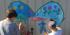 Two kids paint a garden inside the shape of a heart on a fence.