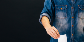 A closeup of someone placing a ballot in a box. The person is wearing a blue denim shirt. The photo is cropped to show only the torso and the hand with the ballot.