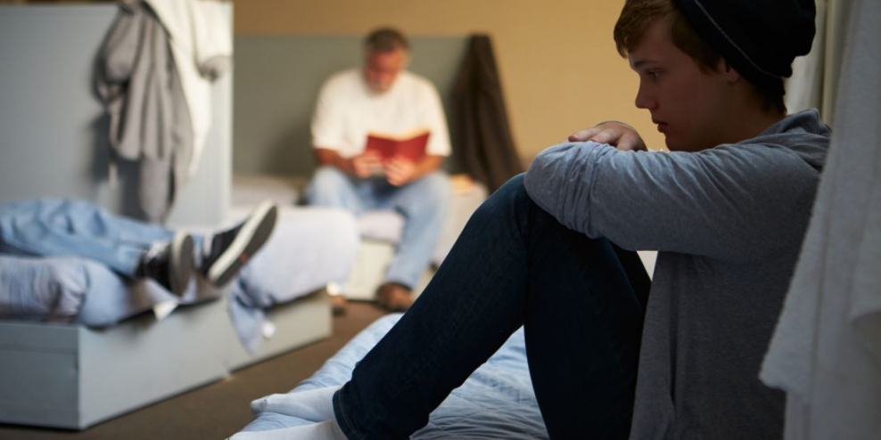 A young person sits against a wall on a bunk with an older man on his own bunk in the background.