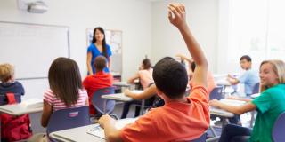 Students in a classroom hold their hands up to answer a question.